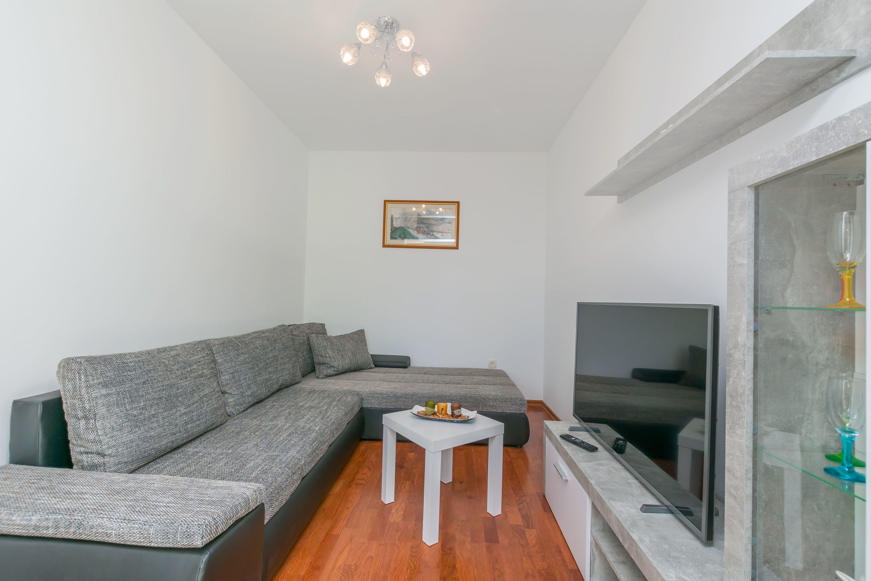 En moderne stue med en grå hjørnesofa, et lille hvidt bord, et fladskærms-tv og et glaskabinet med et vinglas.
