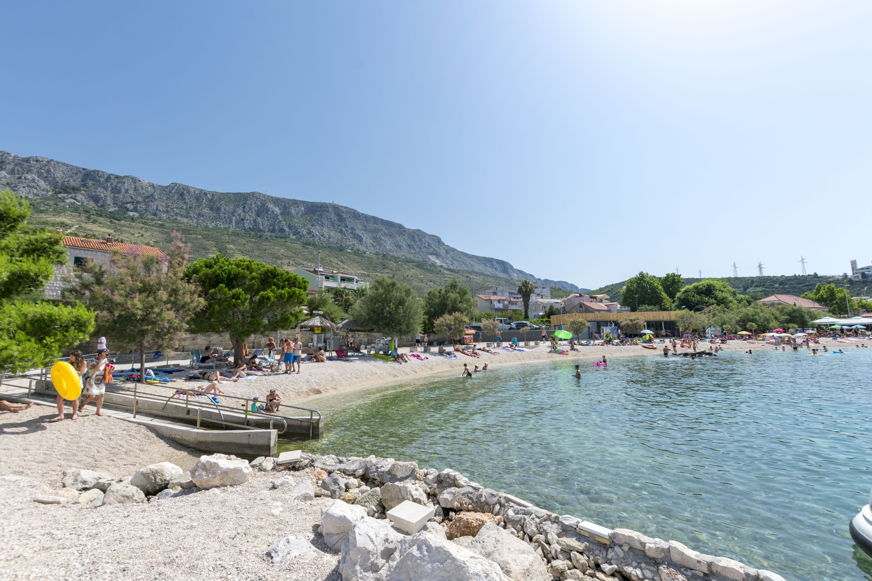 Una playa con aguas cristalinas, costa rocosa y edificios al fondo.