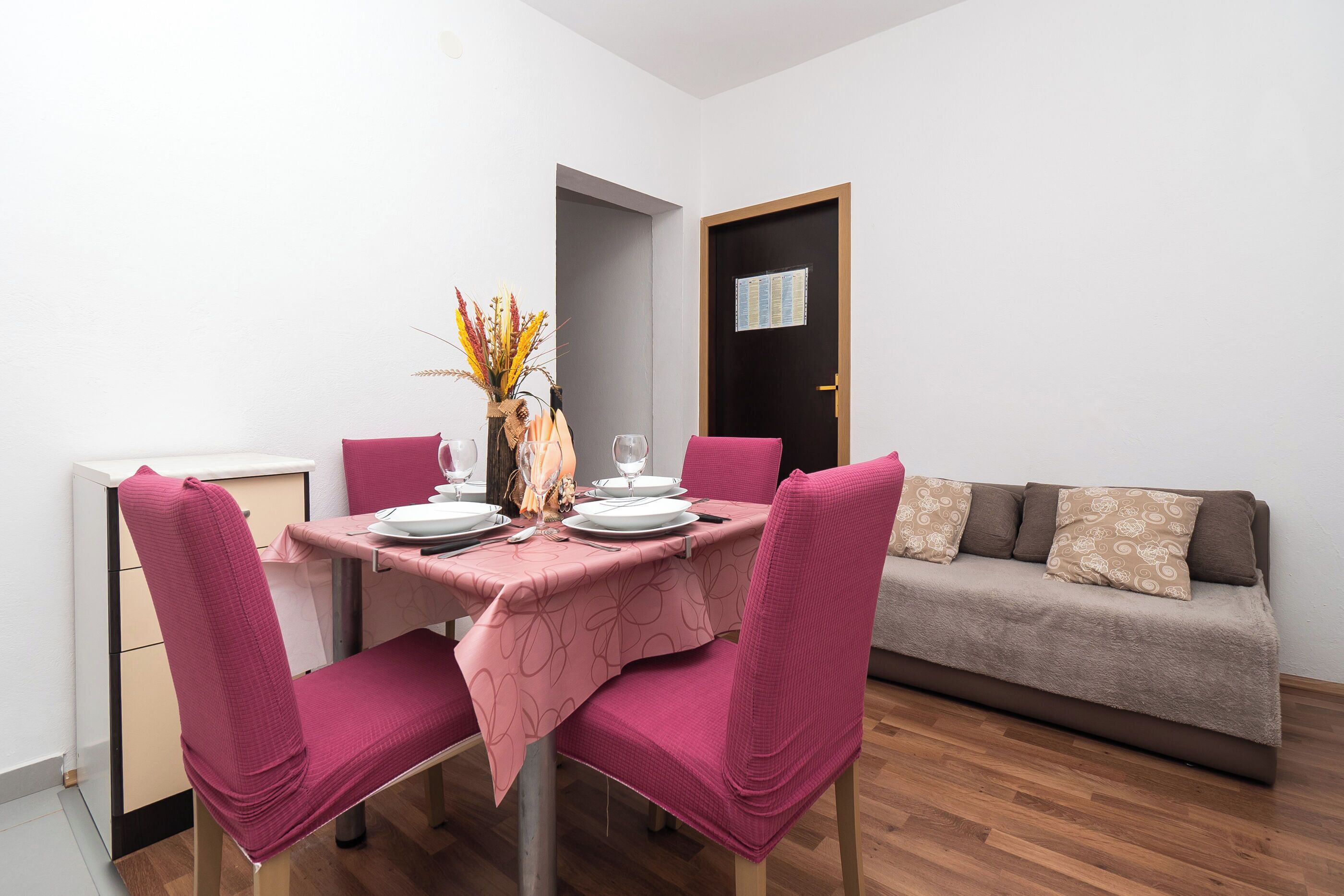 A dining area with a table set for four, pink chairs, a vase with dried flowers, and a sofa in the background.