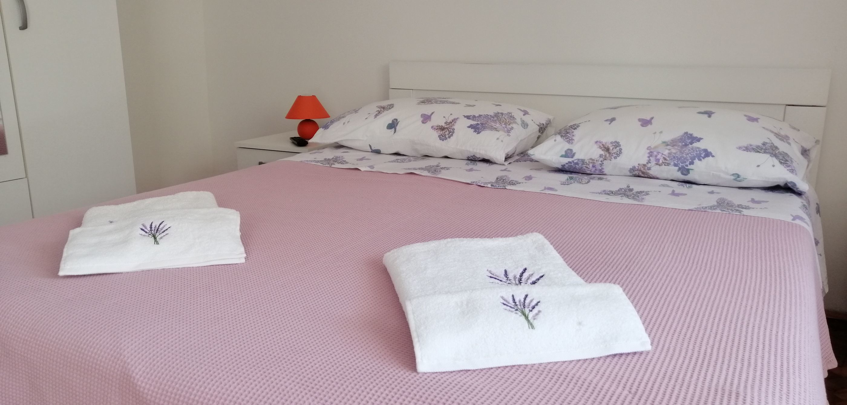Ein ordentlich bezogenes Bett mit blumigen Kissen und einer rosa Tagesdecke.