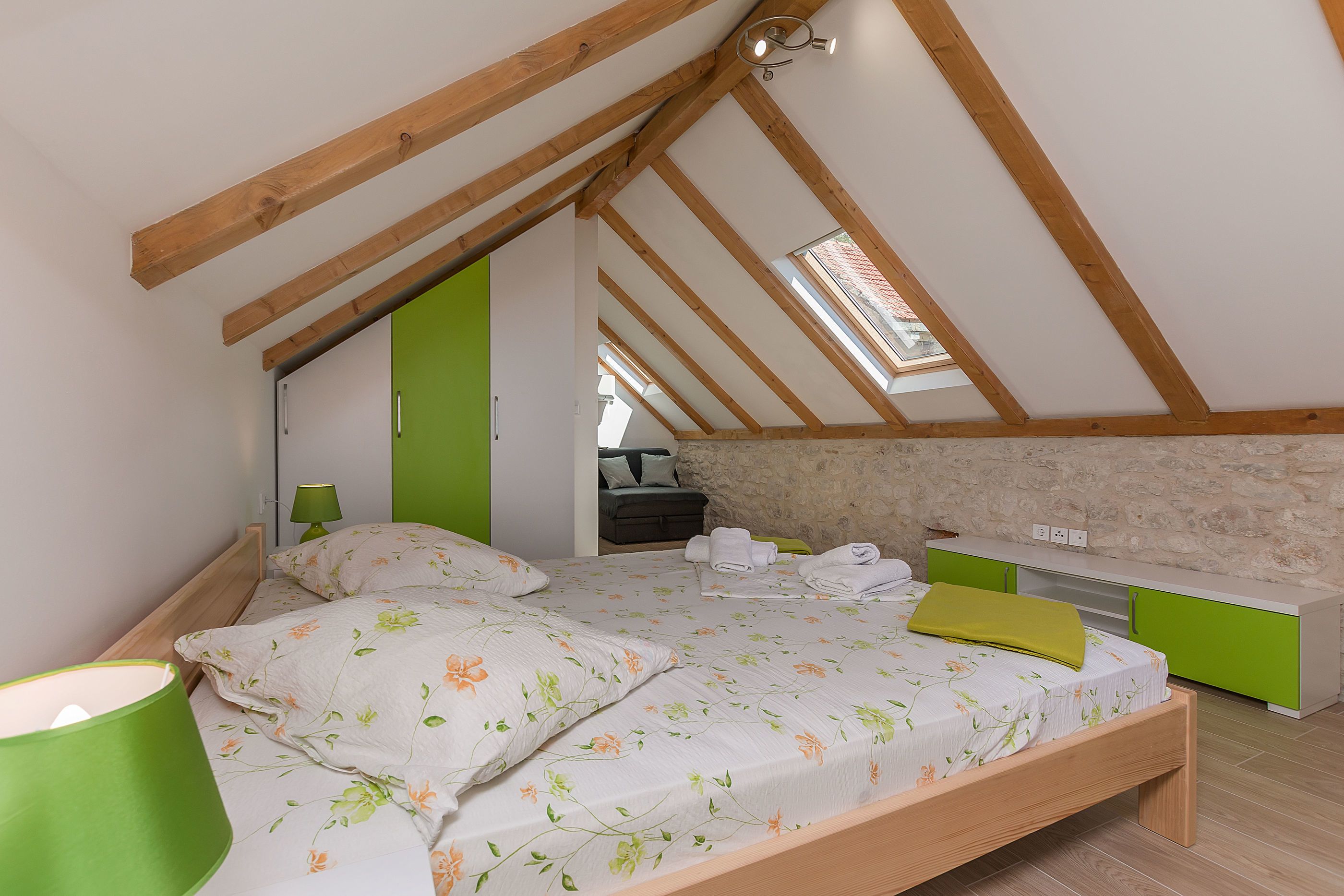 Een slaapkamer met een schuine balklaag, een bed met bloemenbeddengoed, een groen nachtkastje en een bankje met een groen kussen.