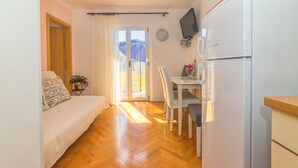 A compact living space with a bed, a dining table with chairs, a refrigerator, a television, and a window with curtains. - Apartments Matko (Podgora)
