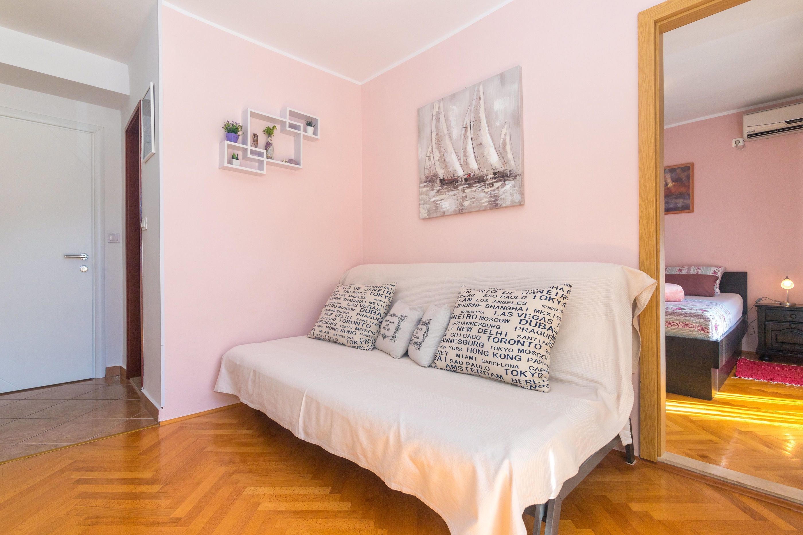 Un dormitorio con cama, pared rosa, pintura de veleros y espejo que refleja otra habitación.