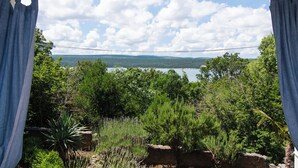 Vista a través de una ventana con cortinas, mostrando un paisaje natural con árboles, una masa de agua y un puente distante.