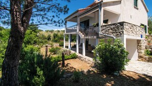 Una casa de dos pisos con un balcón, rodeada de vegetación y un muro de piedra.