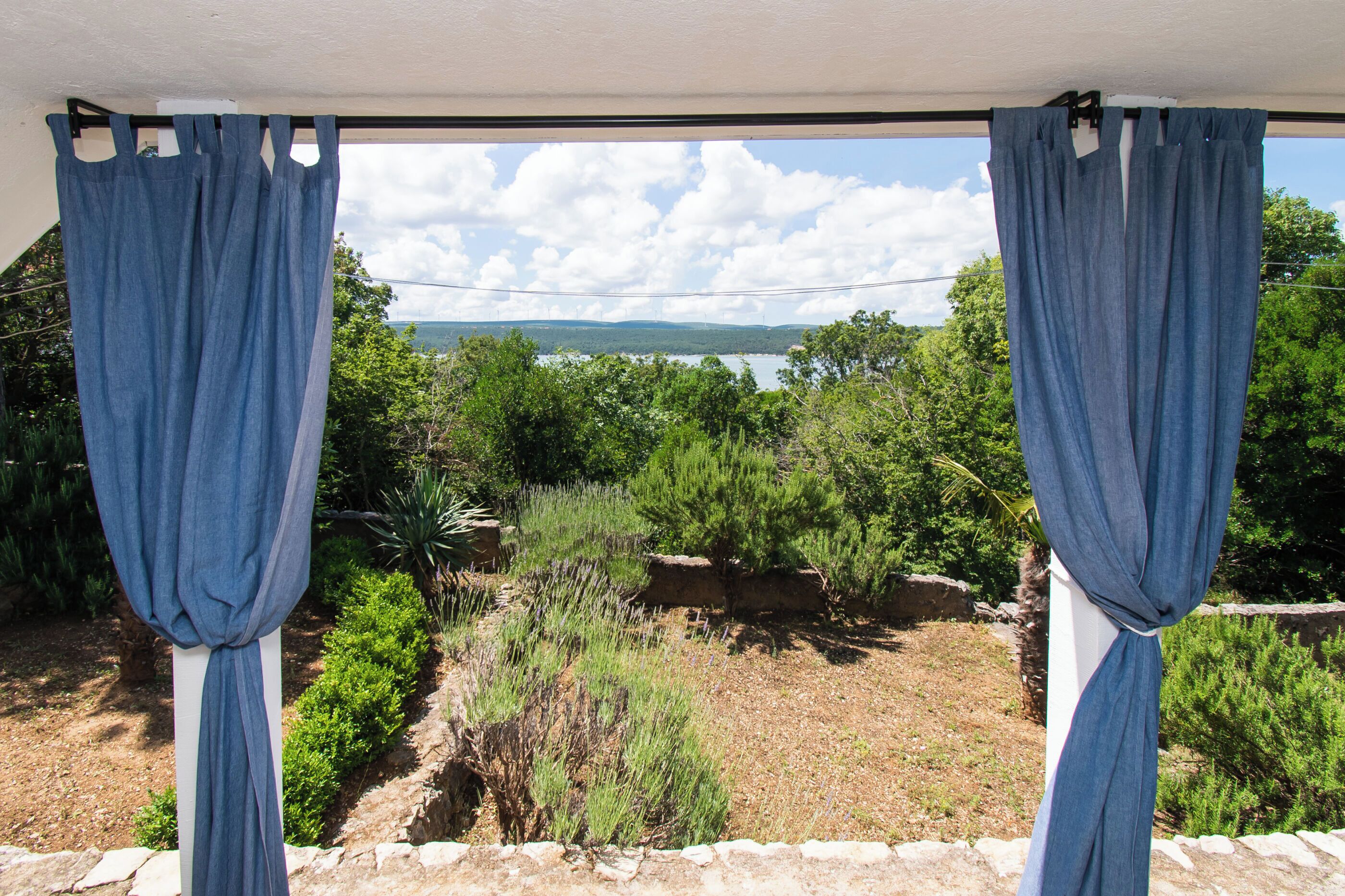 Vista pela janela com cortinas azuis, mostrando uma paisagem com árvores, arbustos e um corpo d'água ao longe.