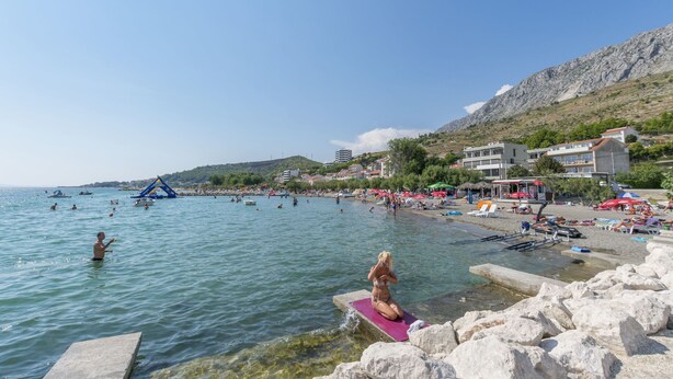 A beach with clear turquoise water, rocky shore, and buildings in the background. - Apartments Villa Aquamarine (Duce)