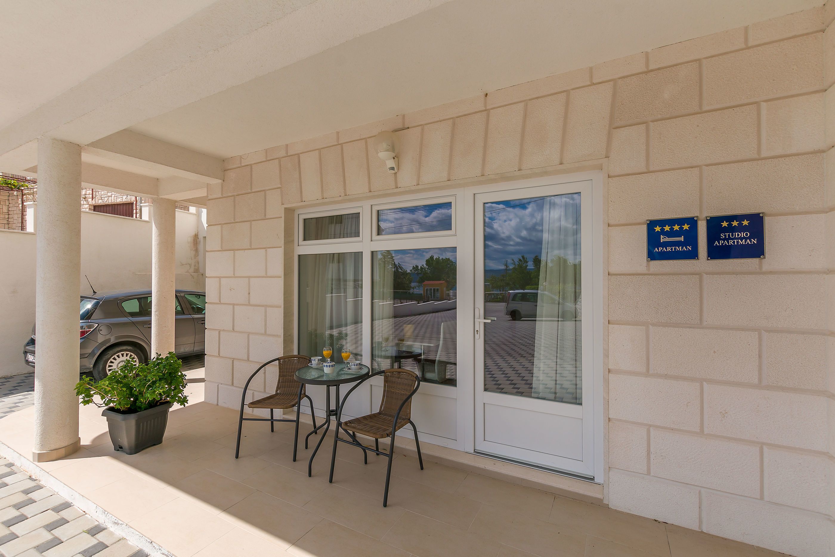 A building with two signs indicating 'APARTMAN' and 'STUDIO APARTMAN'. There is a covered outdoor seating area with chairs and a table, and a potted plant.