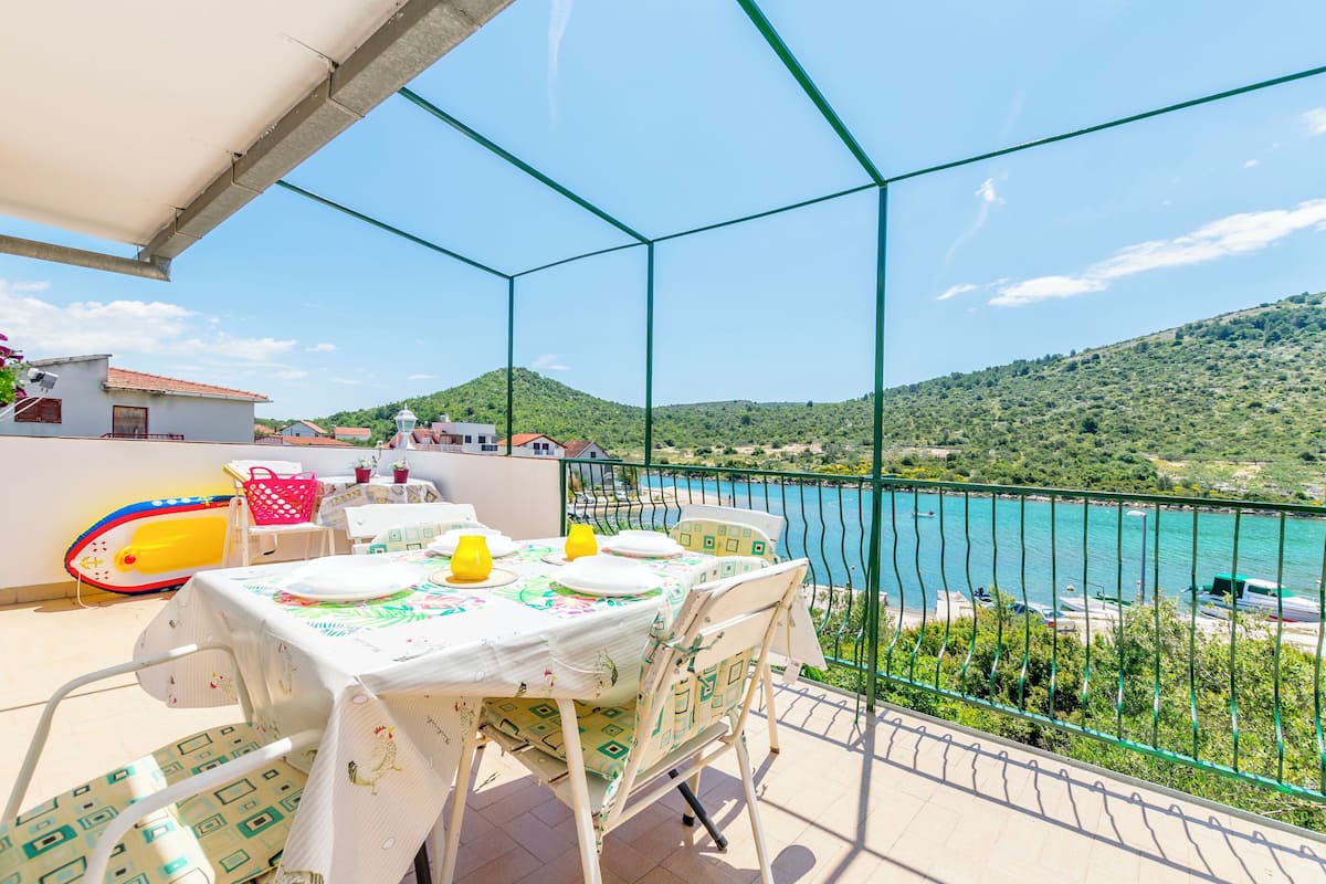 A balcony with a table set for dining, overlooking a turquoise sea and green hills.