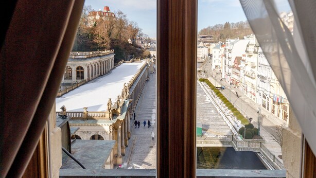 A view through a window with curtains, revealing a snow-covered street and buildings. - Apartments Menuet (Karlovy Vary)