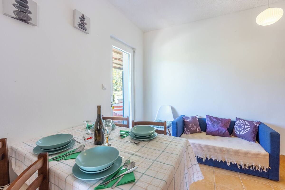 A dining area with a table set for four, a sofa with patterned cushions, and a window with a view of greenery.