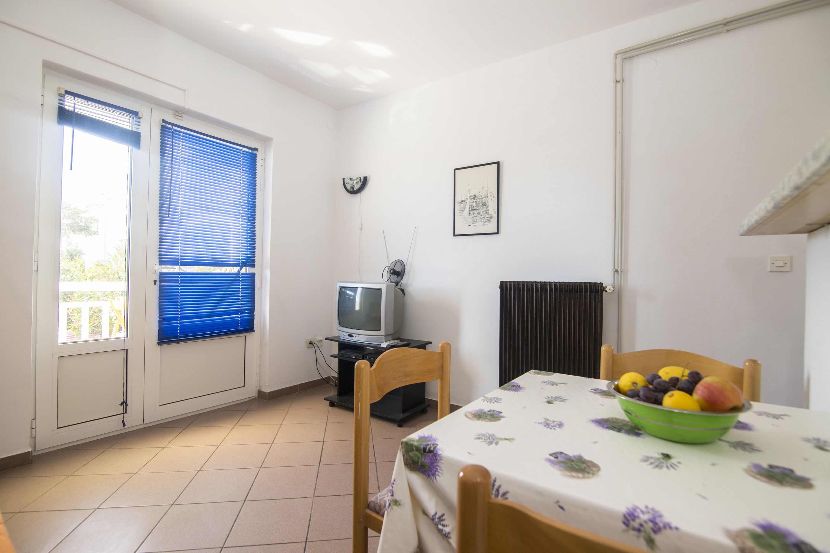 Un coin repas avec une table dressée pour deux personnes, un saladier rempli de fruits, un téléviseur posé sur un meuble et une fenêtre avec des stores.