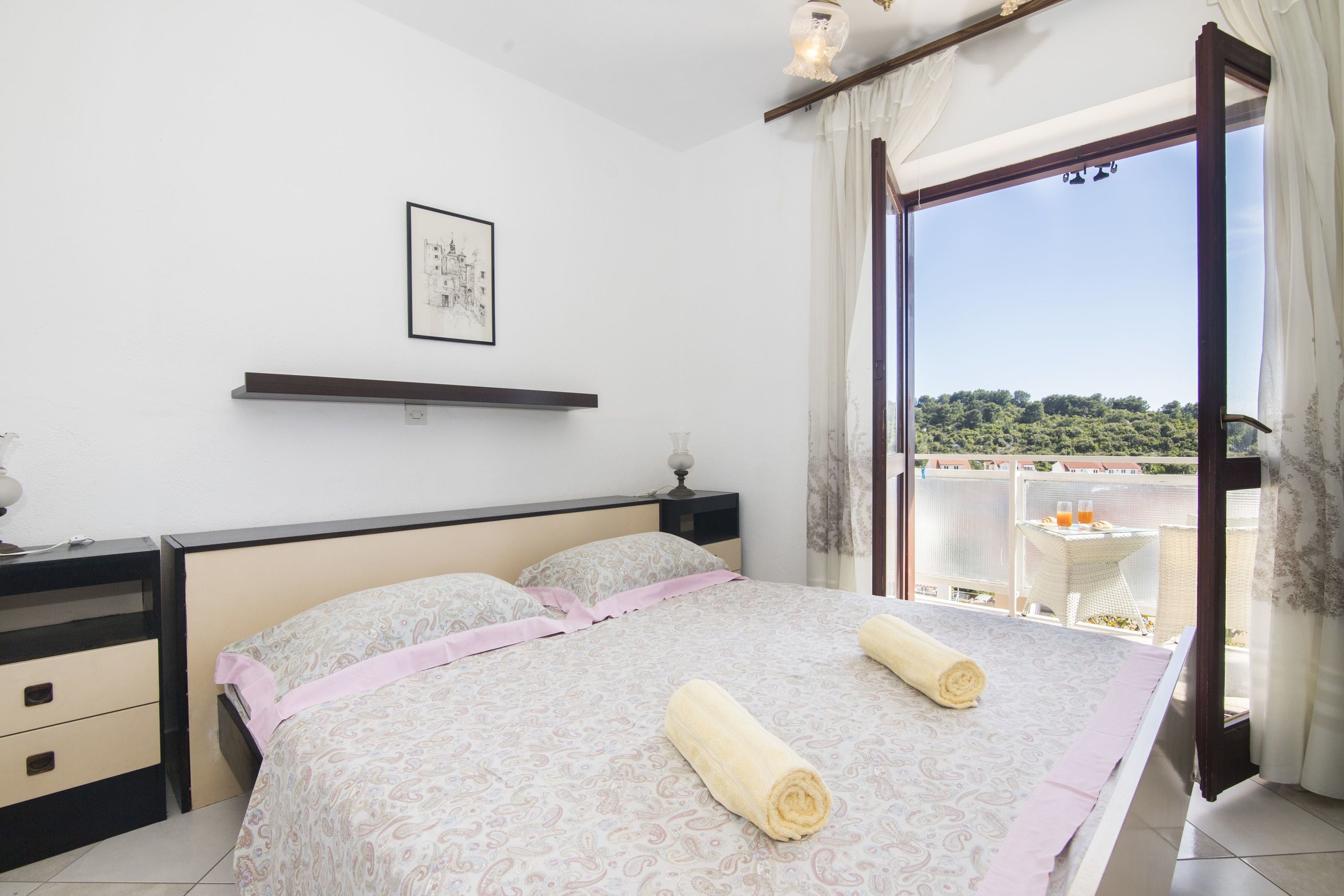 Un dormitorio con una cama grande, una mesita de noche, una lámpara de noche, un cuadro en la pared y vistas al exterior a través de un ventanal amplio.