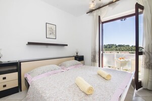 Un dormitorio con una cama grande, una mesita de noche, una lámpara de noche, un cuadro en la pared y vistas al exterior a través de un ventanal amplio.