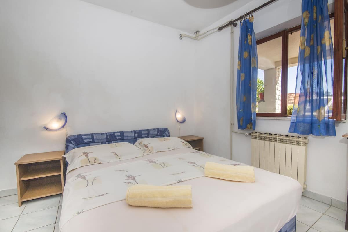 Une chambre à coucher avec un lit, deux tables de chevet, une fenêtre avec des rideaux bleus et un luminaire fixé au mur.