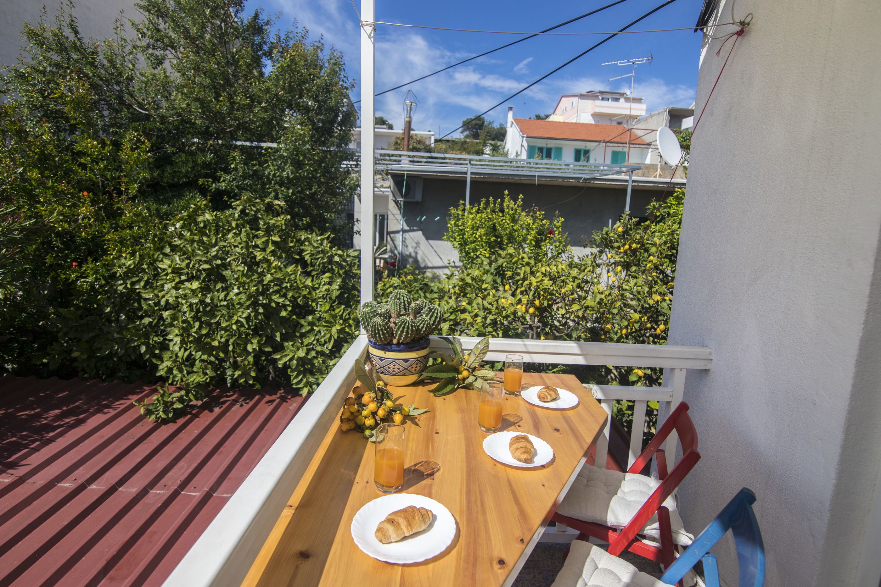 A rooftop terrace with a wooden table set for a meal, surrounded by greenery and a cityscape in the background.