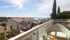 A balcony with a wicker chair, a table with a glass of orange juice and a plate of food, and a view of a coastal town with buildings and trees. - Apartments and Rooms Nikol (Hvar)
