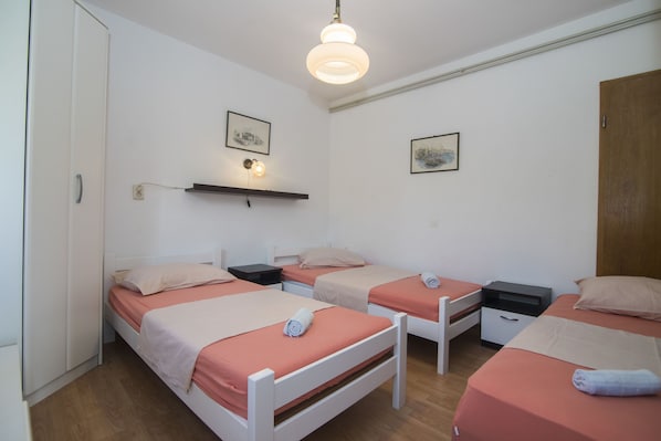 Una habitación con tres camas individuales, cada una con estructura blanca y ropa de cama naranja. Hay mesitas de noche con lámparas, un estante en la pared sobre las camas y una puerta de madera a la derecha.