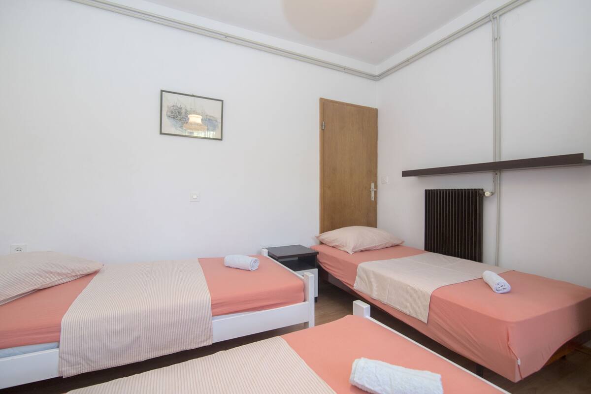 Une chambre avec trois lits simples, chacun recouvert d’une couverture rose et agrémenté d’oreillers blancs. On y trouve une porte en bois et un radiateur au mur.