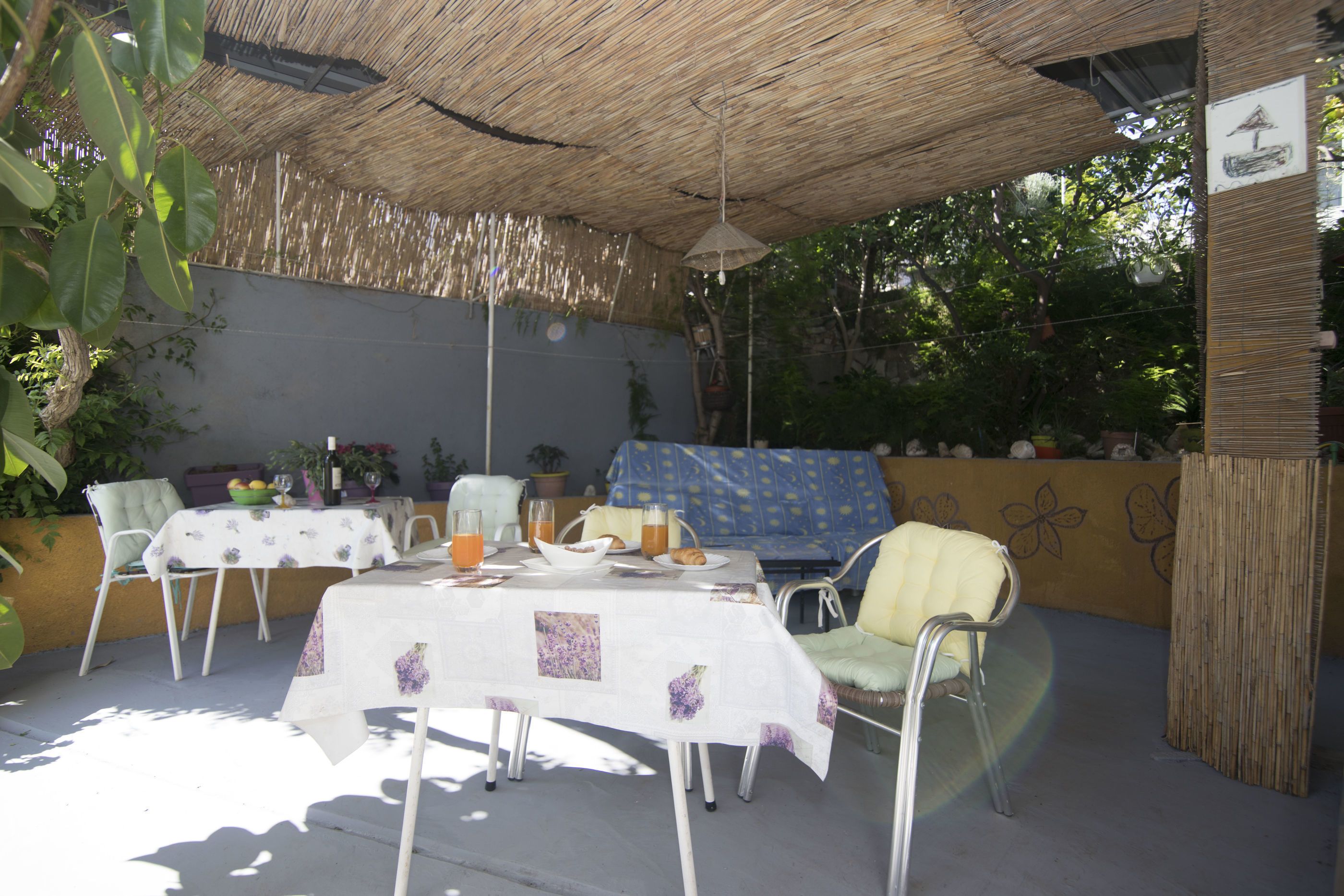 Un espace extérieur aménagé pour les repas, avec des tables et des chaises, abrité par un toit de chaume et entouré de plantes en pot et d’un mur décoratif.