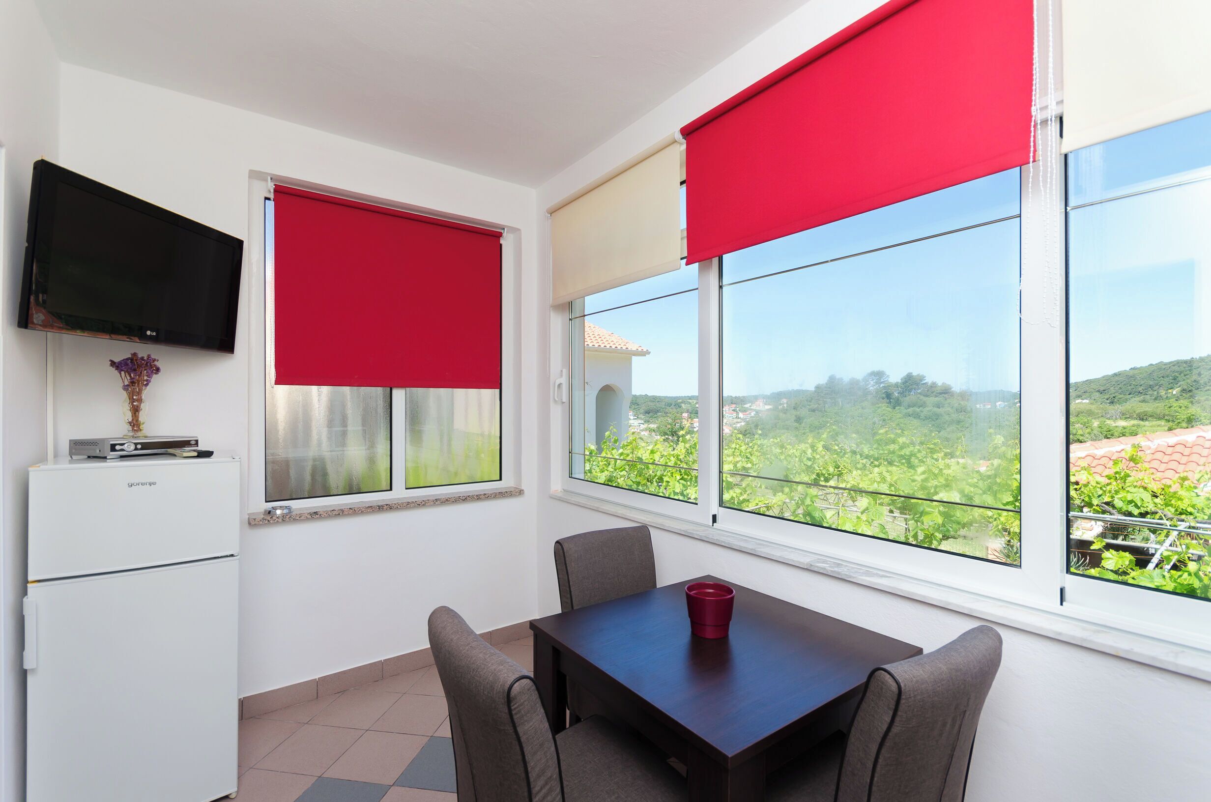 A room with a dining table, chairs, a TV mounted on the wall, and large windows with red and beige blinds.