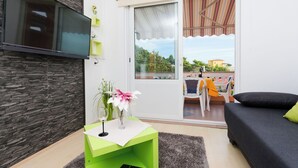 Ein modernes Wohnzimmer mit einem Flachbildfernseher an einer strukturierten Wand, einem grünen Beistelltisch mit Vase und Fernbedienung, einem Sofa mit einem grünen Kissen sowie einem Balkon mit Tisch und Stühlen.
