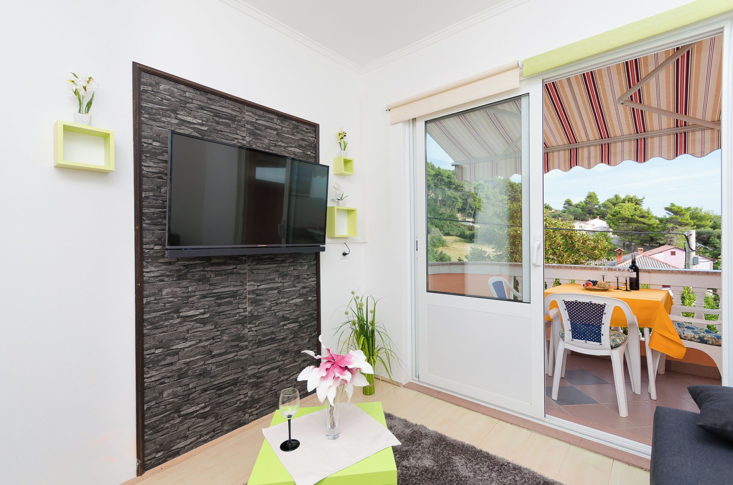 Una sala de estar moderna con un televisor de pantalla plana montado en una pared de aspecto pétreo, un comedor con mesa y sillas, y un puerta corrediza de vidrio que da a un balcón exterior.