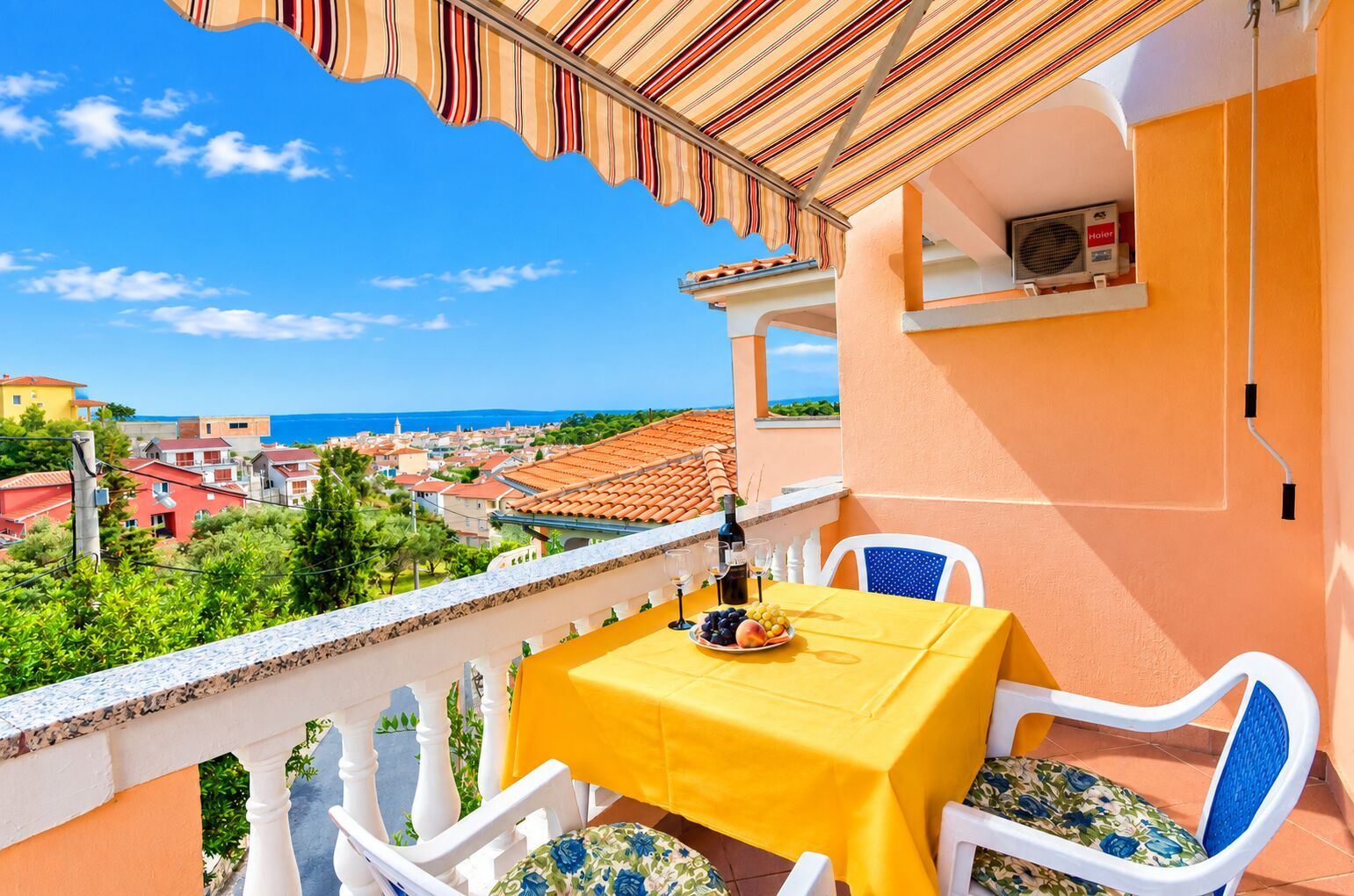 Ein Balkon mit einem gelben Tisch, weißen Stühlen und Blick auf das Meer und die Stadt.