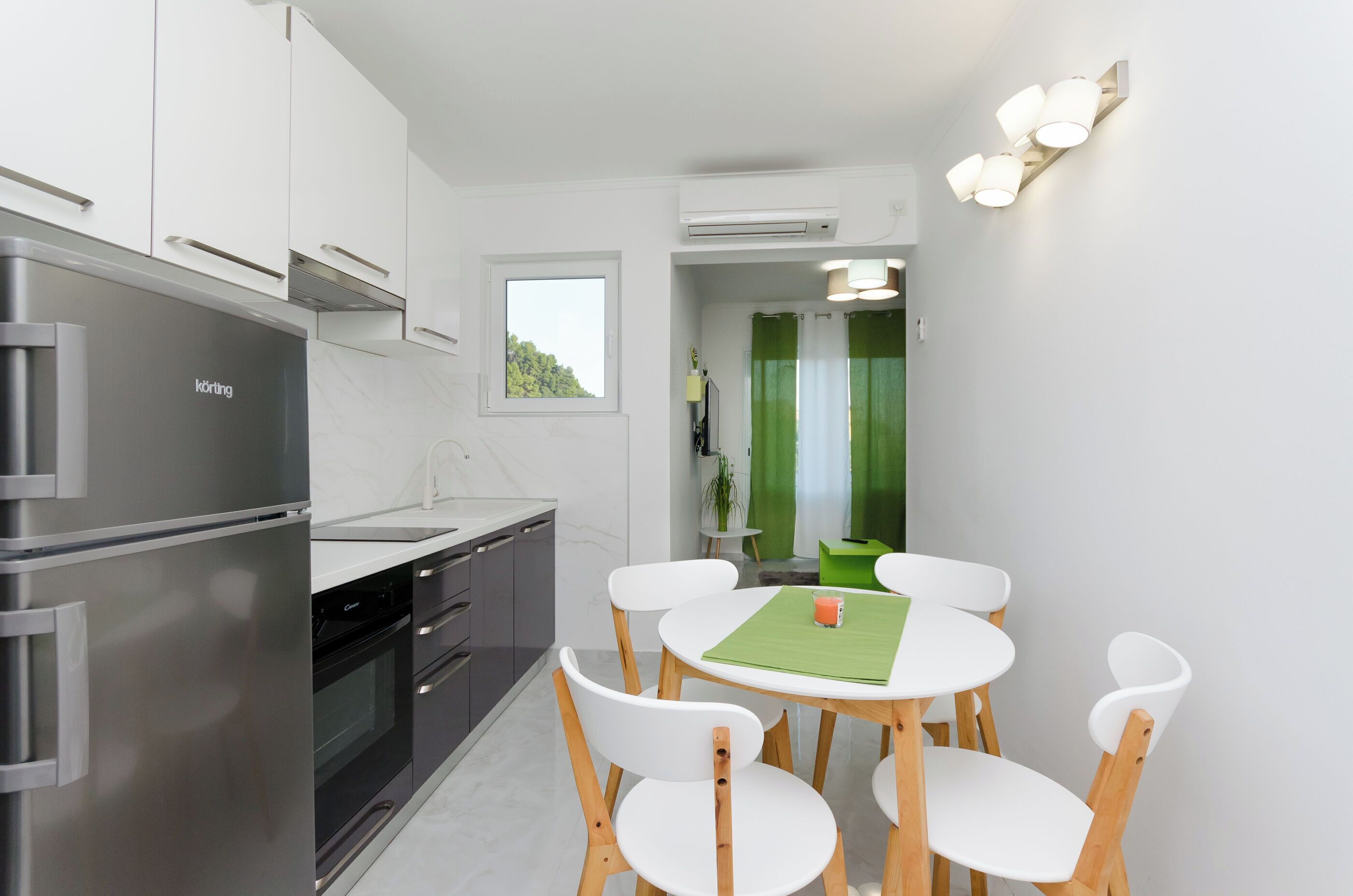 A modern kitchen with a stainless steel refrigerator, built-in oven, and white cabinetry. There is a dining area with a round table and white chairs.