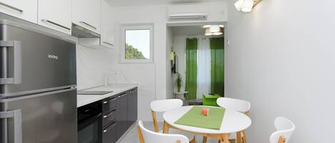 A modern kitchen with a stainless steel refrigerator, built-in oven, and white cabinetry. There is a dining area with a round table and white chairs.