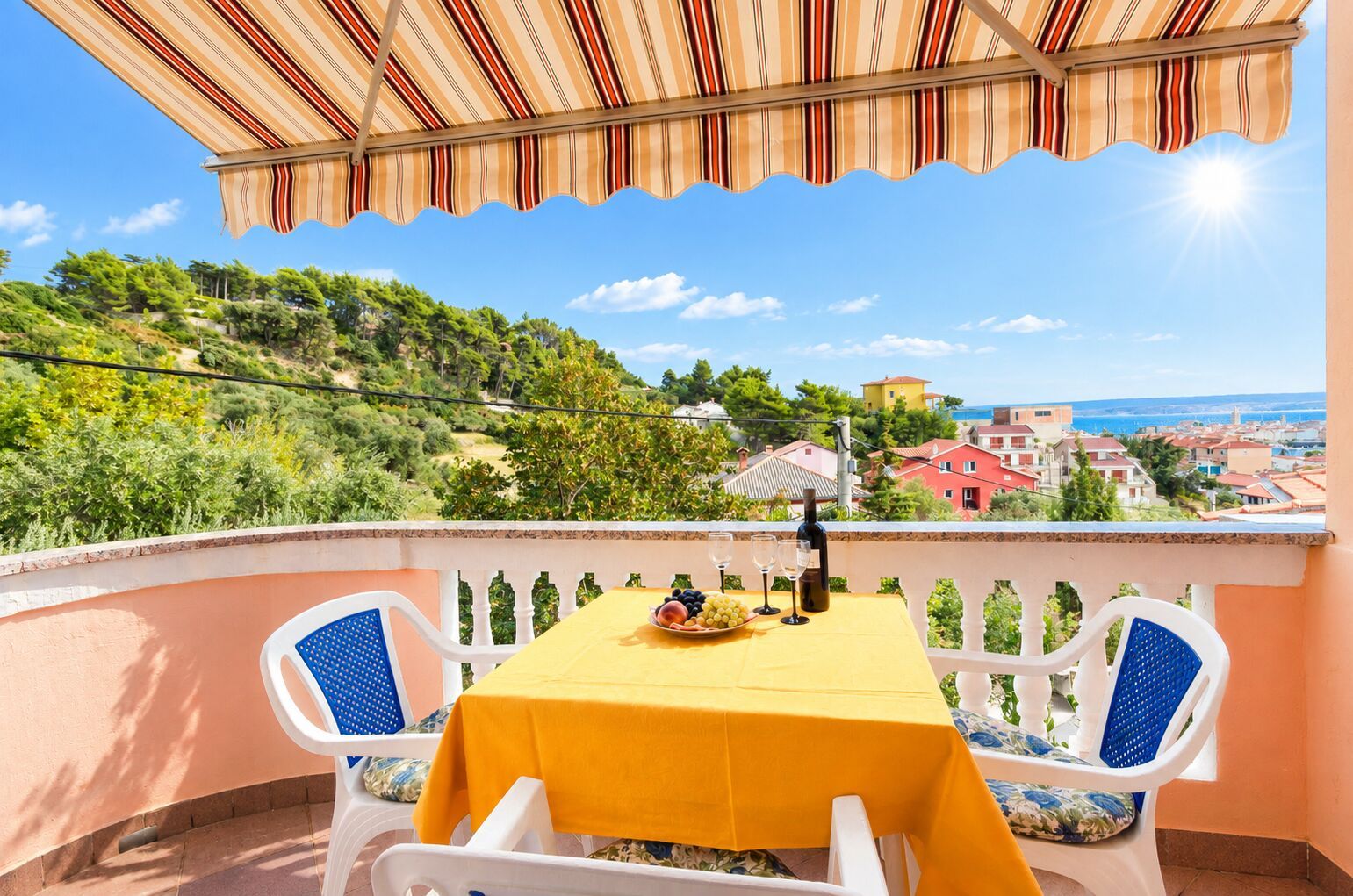 A balcony with a yellow table, two chairs, a bottle, and a bowl of fruit.
