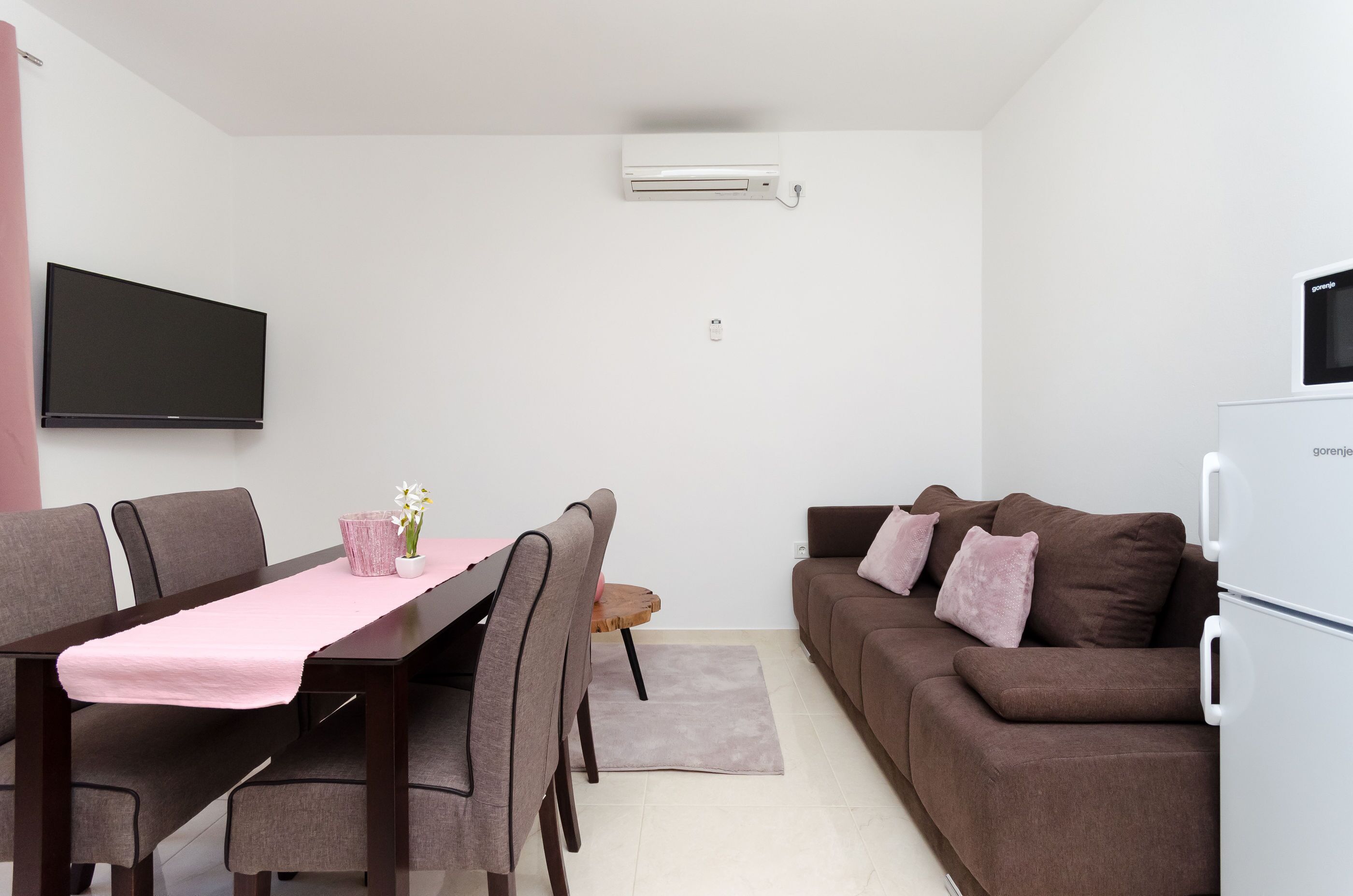 Ein modernes Wohnzimmer mit Essbereich, einem wandmontierten Flachbildfernseher, einem weißen Kühlschrank und einem braunen Sofa mit Kissen.