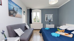 A modern bedroom with a grey sofa, a bed with blue bedding, a bedside table with a vase and a tray of breakfast items, and a window with a view of greenery. - Apartments Ester (Funtana)