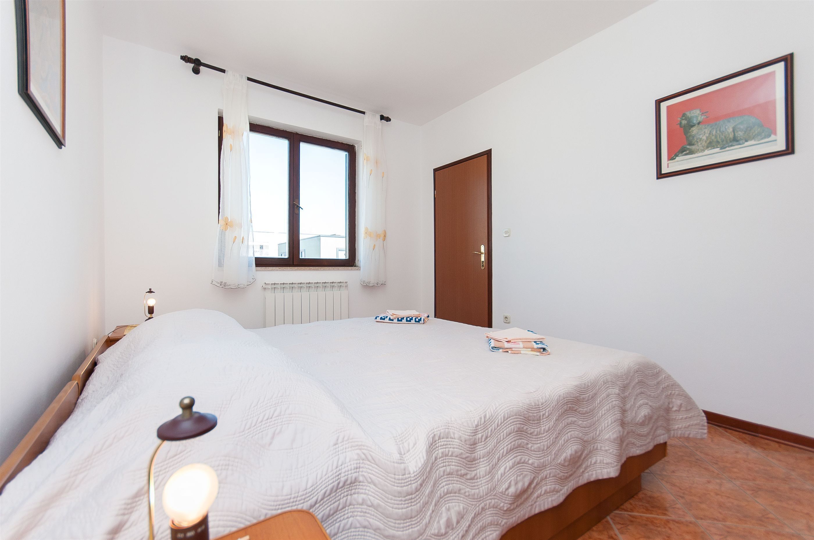 Ein Schlafzimmer mit Bett, Fenster mit Vorhängen, Tür, einem gerahmten Bild an der Wand und einem Nachttisch mit Lampe.