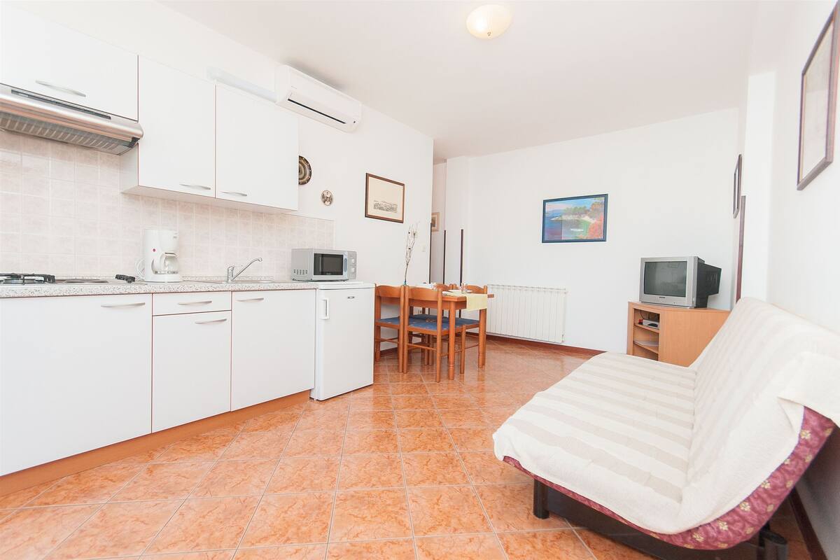 A compact kitchen with white cabinets, a microwave, and a coffee maker. There is a dining table with chairs and a TV on a wooden stand.