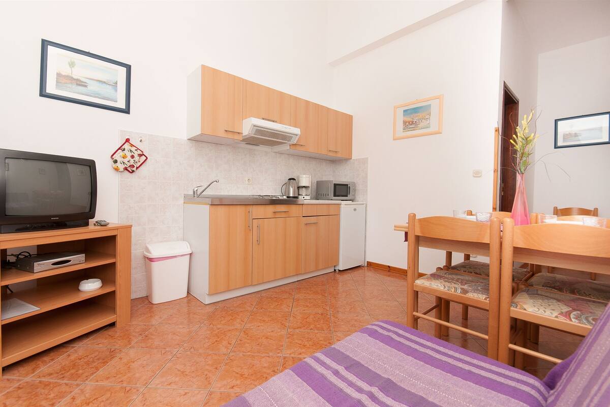 A compact kitchen with wooden cabinets, a microwave, and a coffee maker. There is a television on a stand, a trash bin, and a dining table with chairs.