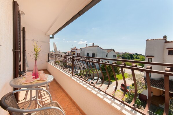 En balkon med et bord og stole, en vase med blomster, og udsigt til omkringliggende bygninger og en gade.