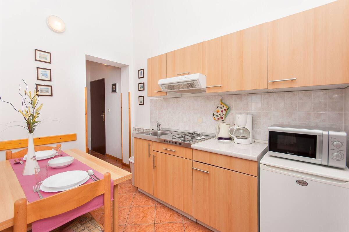 A compact kitchen with wooden cabinets, a microwave, a coffee maker, and a dining area with a pink tablecloth.