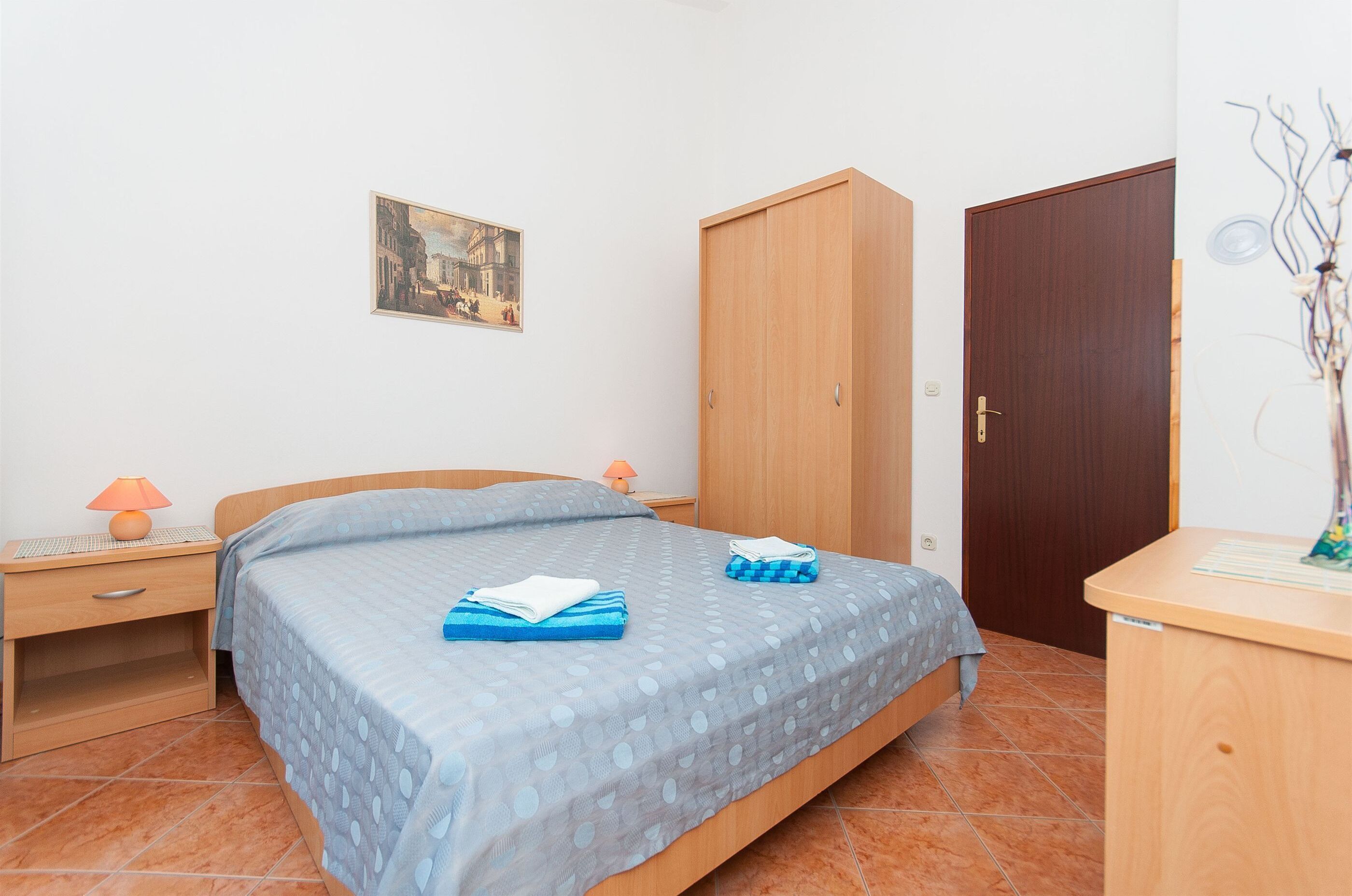 A bedroom with a bed, a wooden wardrobe, a nightstand with a lamp, and a painting on the wall.