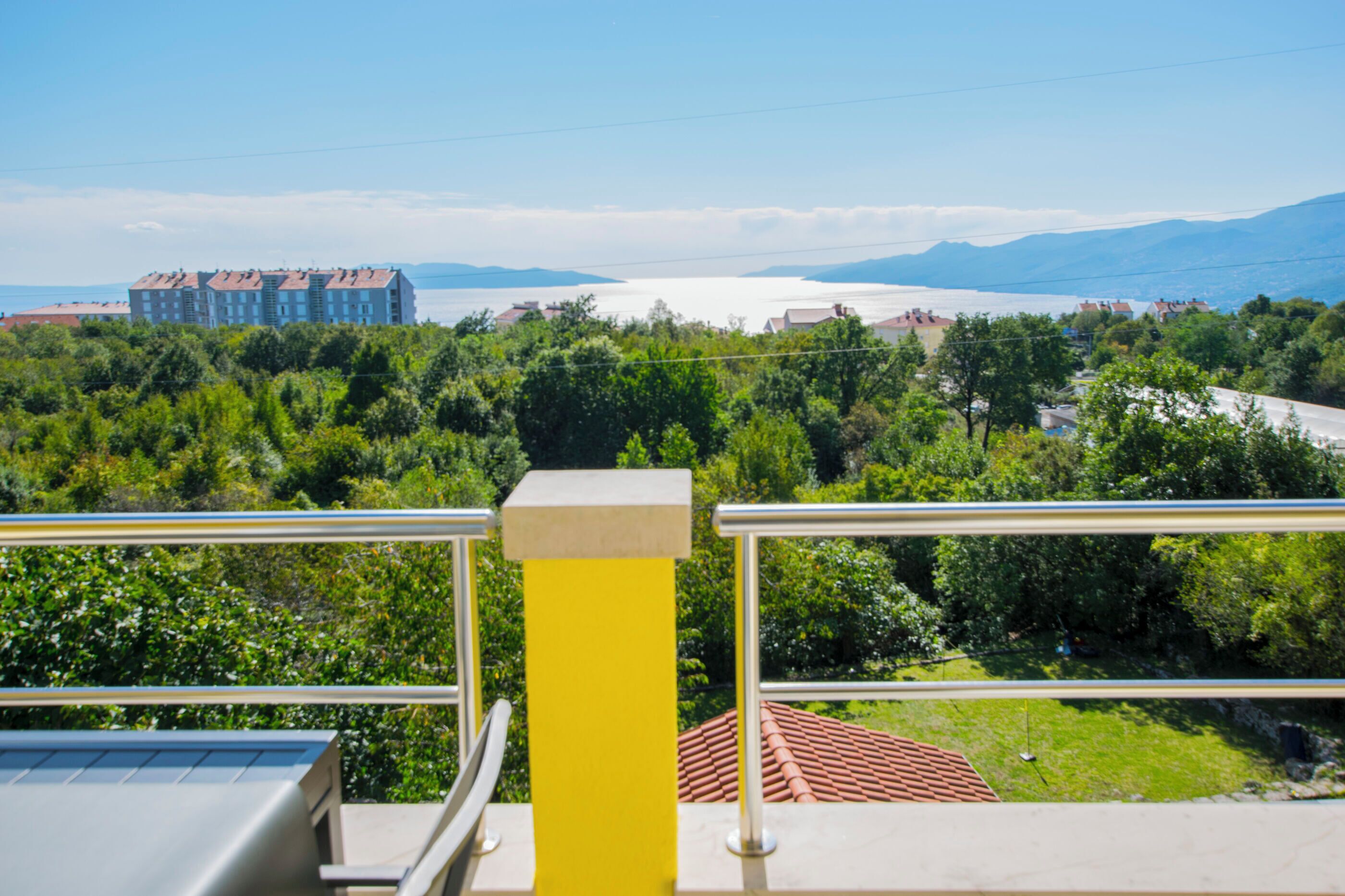 A balcony with a yellow railing overlooking a coastal area with buildings and greenery.