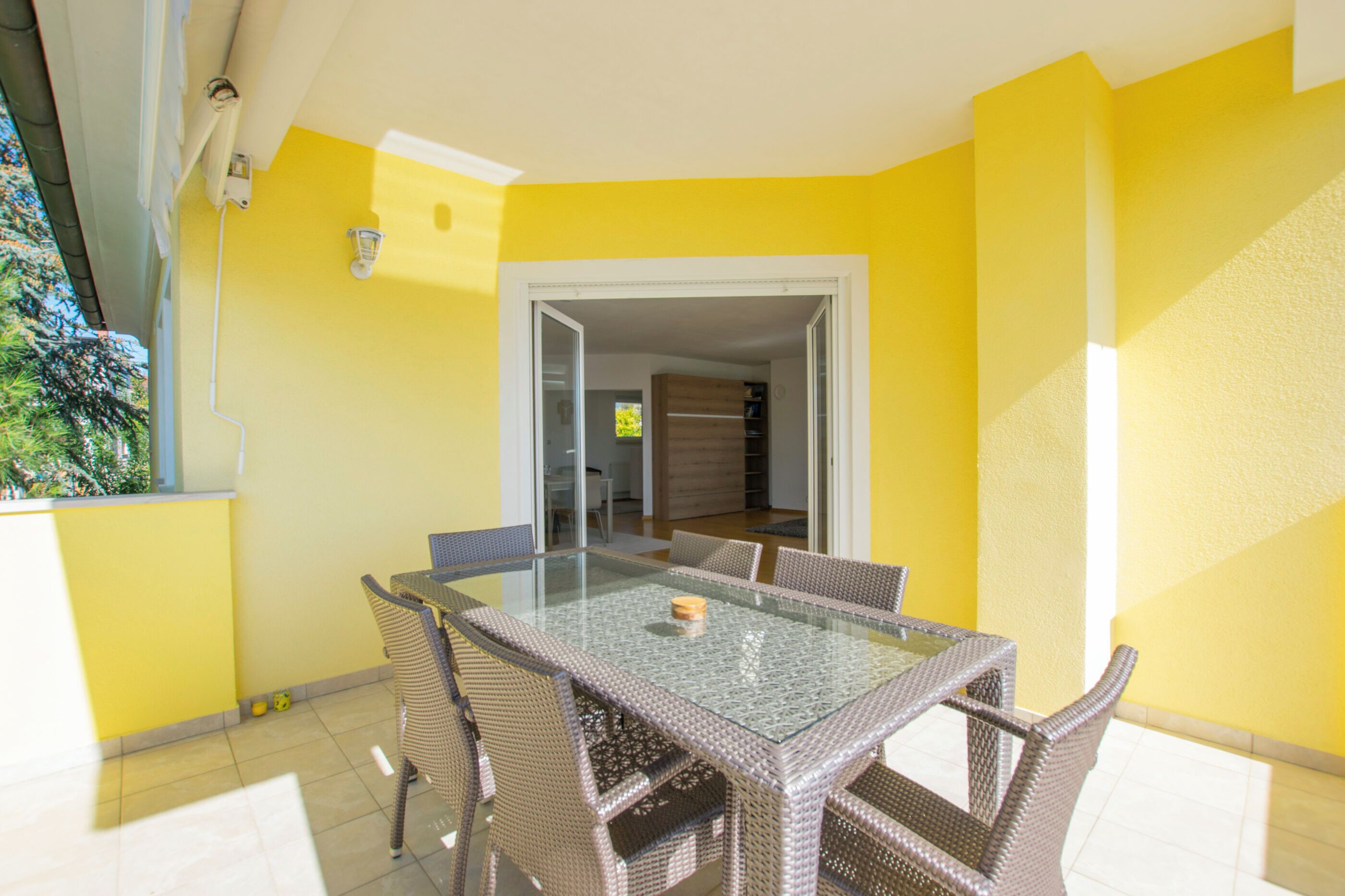 A dining area with a glass table and wicker chairs, yellow walls, and a view of the interior through an open door.