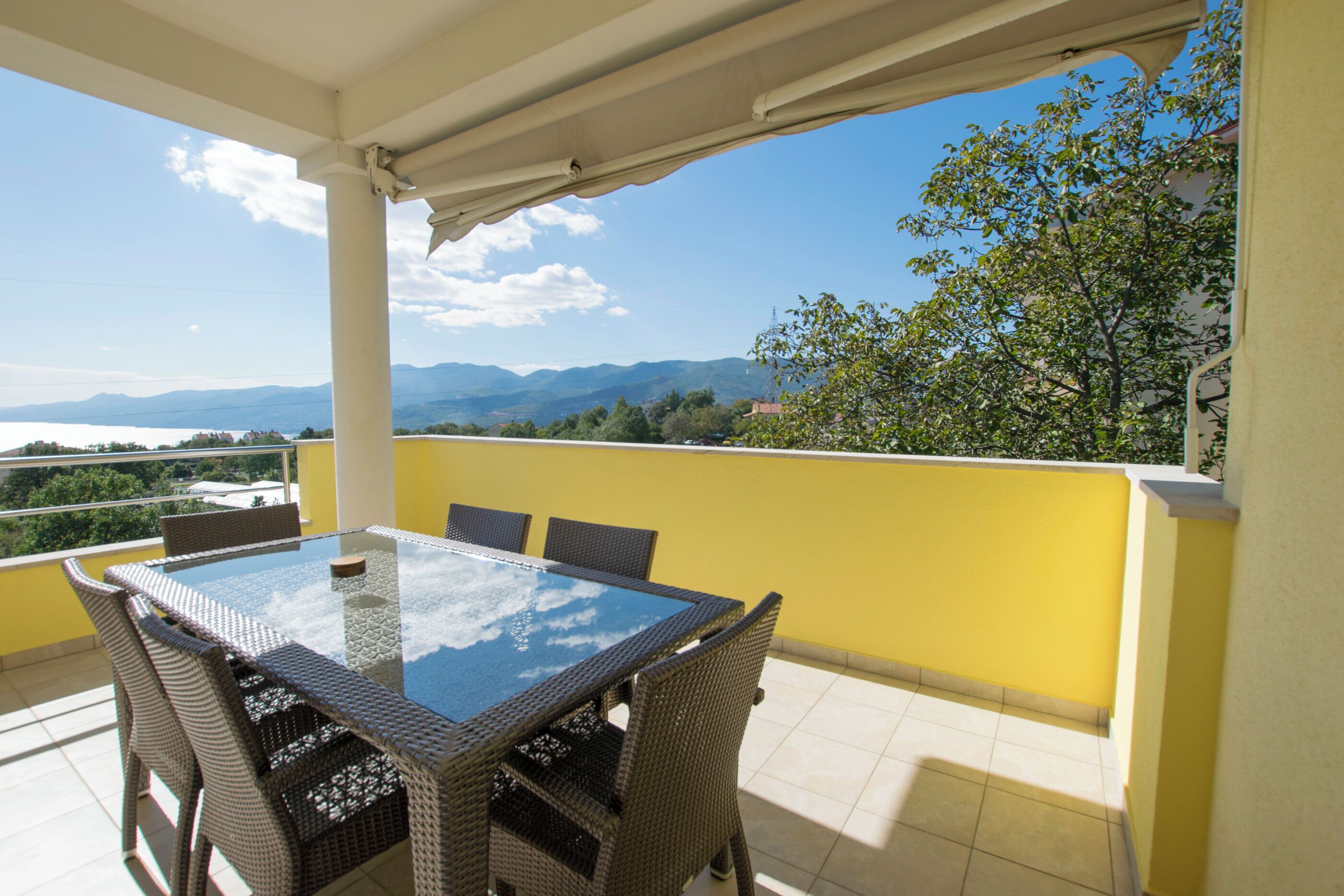Un balcón con una mesa de vidrio y sillas de mimbre, con vistas a un paisaje montañoso.