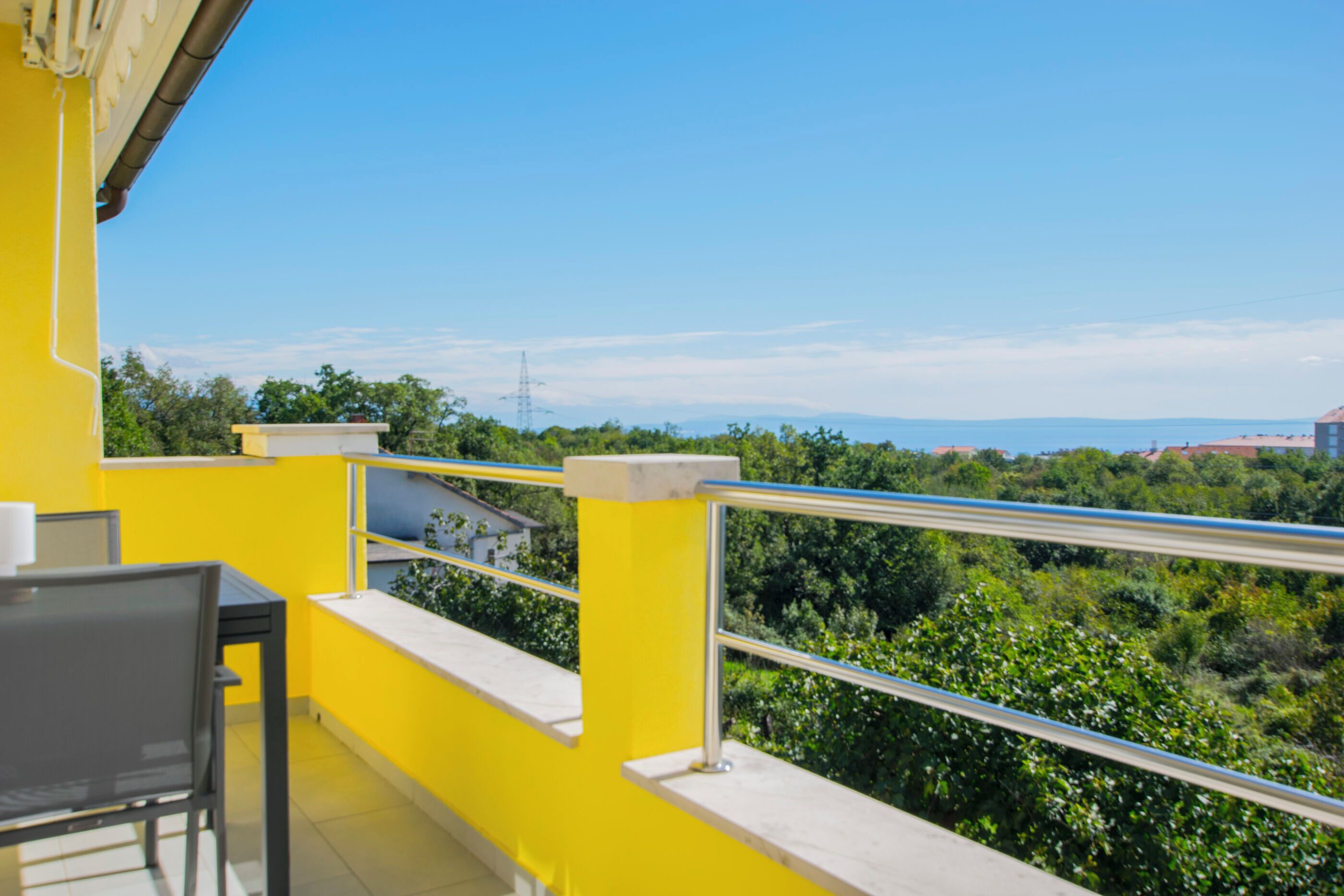 Un balcone con ringhiere gialle, un tavolo e una sedia, affacciato su un paesaggio verde e lussureggiante e edifici in lontananza.