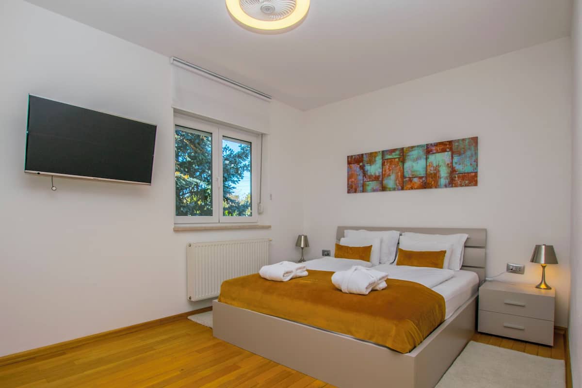 A modern bedroom with a large bed, a flat-screen TV, a window with a view of trees, and a painting on the wall.