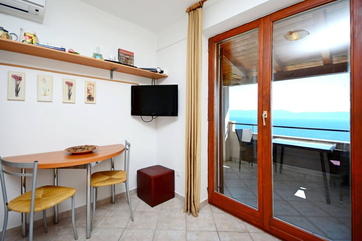 Une pièce avec une table et des chaises en bois, un téléviseur fixé au mur et une porte-fenêtre coulissante donnant sur un espace extérieur avec vue sur la mer.