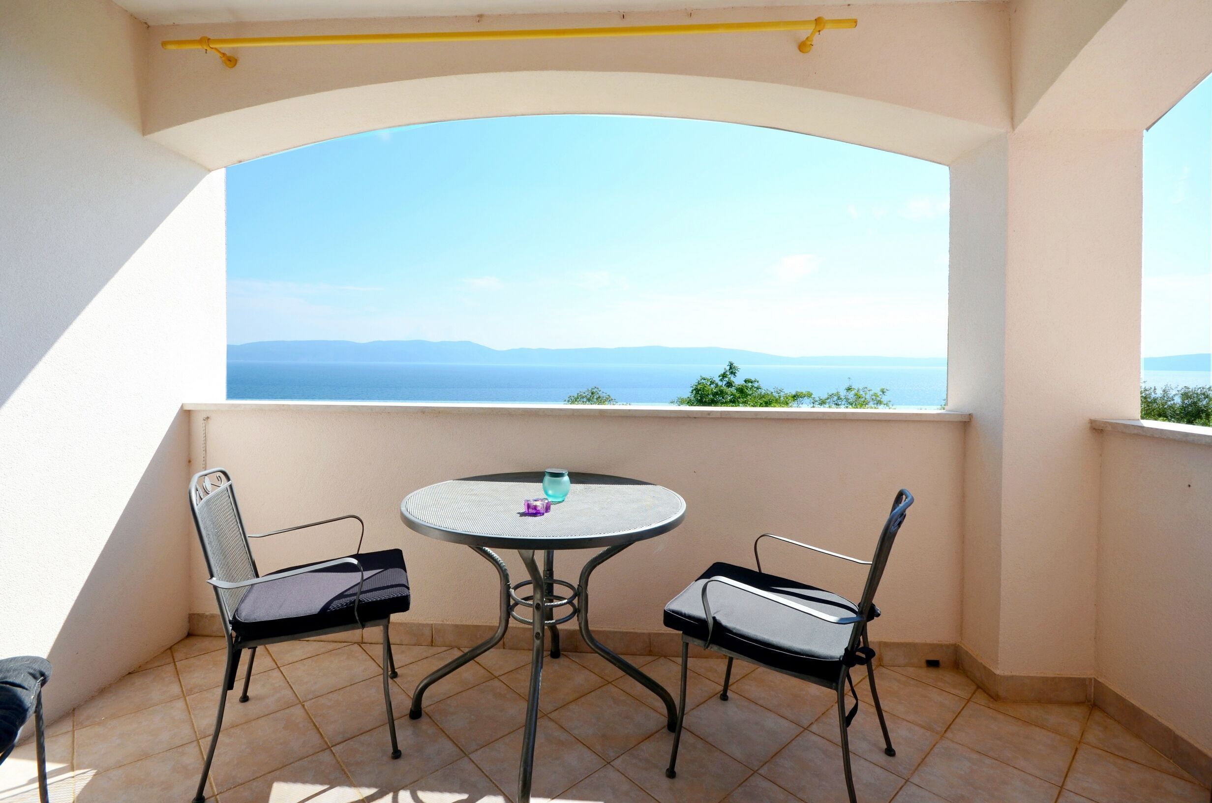 A balcony with a round table and two chairs, overlooking a scenic view of the sea and distant mountains.