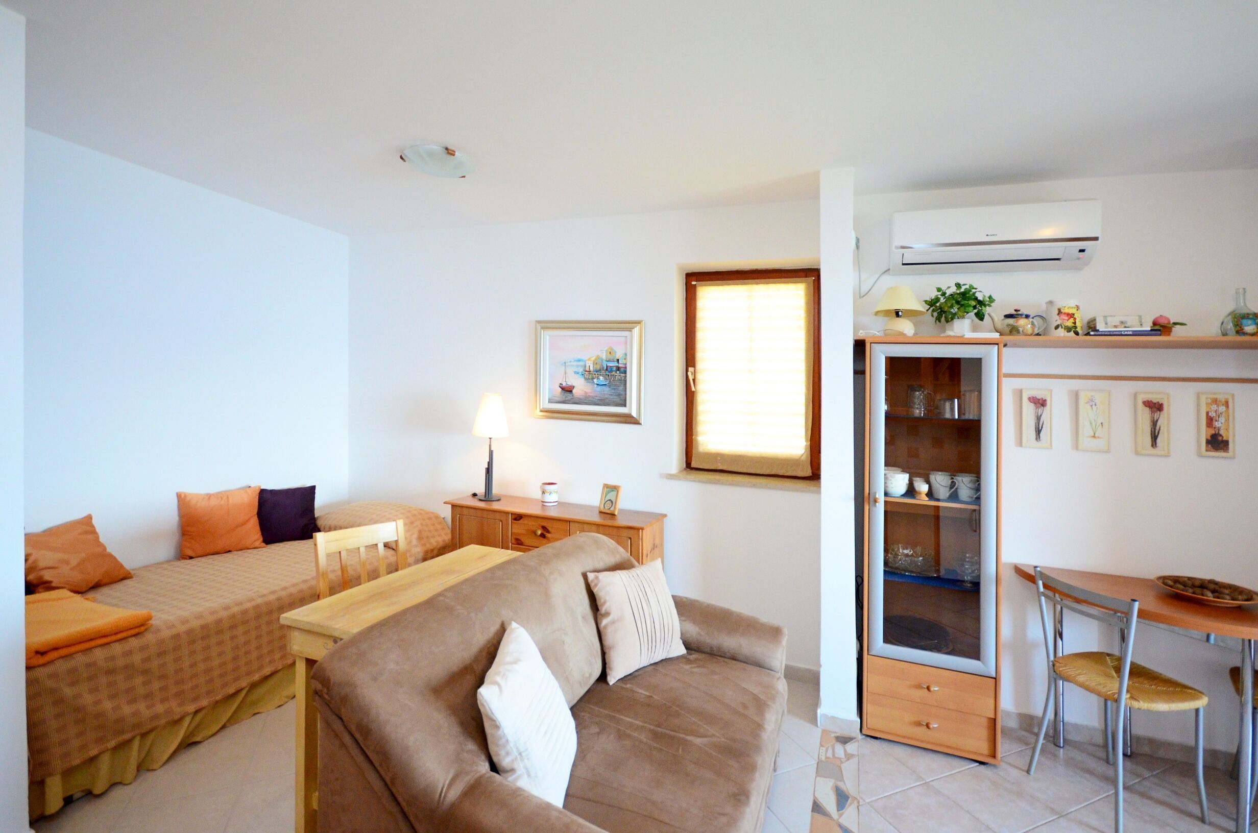 A compact living space with a sofa, a small dining table, a cabinet with glass doors, and a wall-mounted air conditioner.