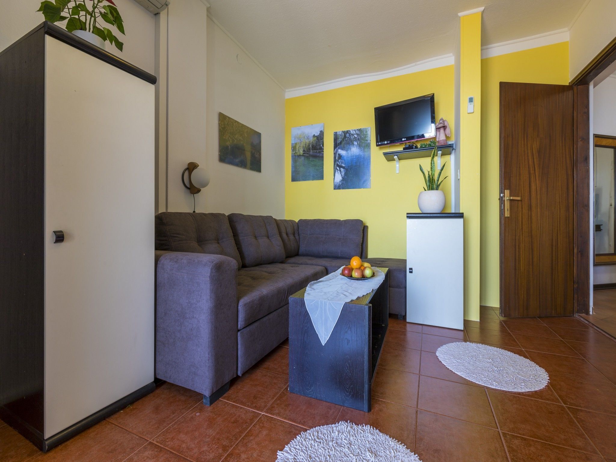 Una sala de estar con un sofá gris, una mesita de centro con fruta, un televisor sobre un mueble bajo y una puerta a la derecha.