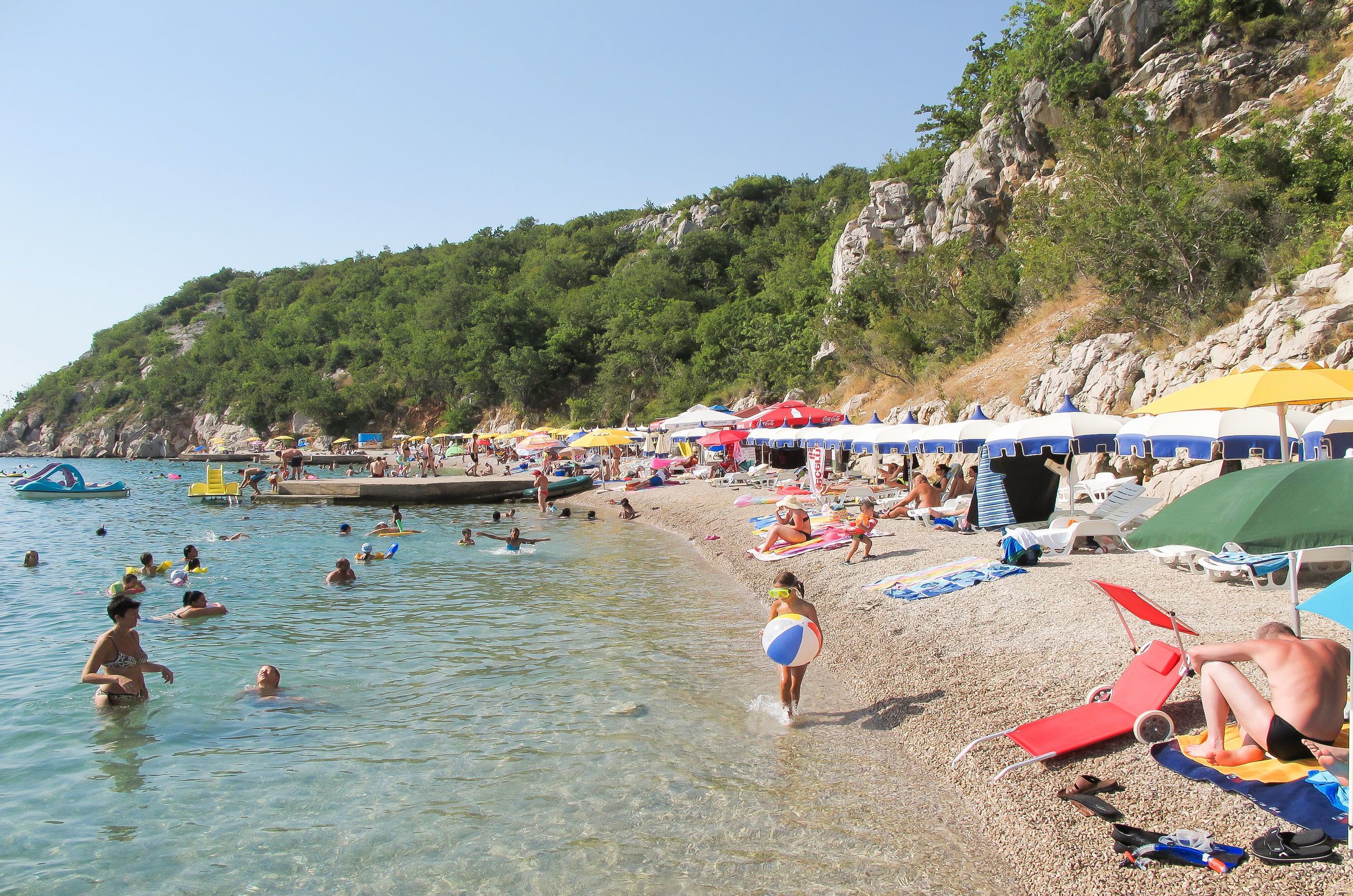 Ein Strand mit klarem Wasser, Menschen beim Schwimmen und Sonnenbaden, und Sonnenschirme spenden Schatten.