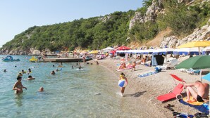 Ein Strand mit klarem Wasser, Menschen beim Schwimmen und Sonnenbaden, und Sonnenschirme spenden Schatten.